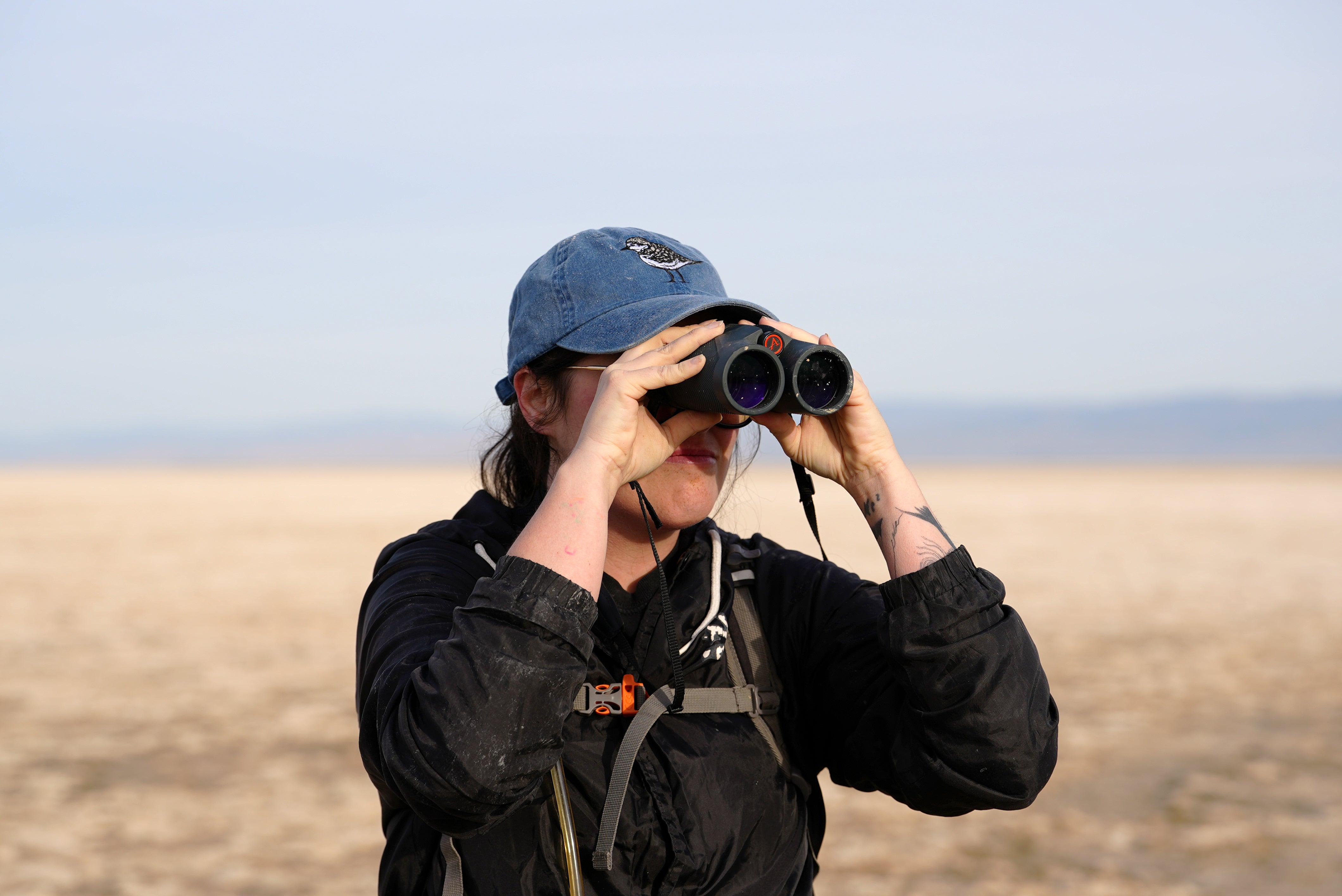 A woman in a black jacket and a blue cap embroidered with a Snowy Plover uses binoculars to scan the horizon at the Salton Sea. They have a backpack with a hydration tube and visible tattoos on their arm. The background features a vast, dry landscape with